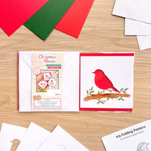 Load image into Gallery viewer, A Christmas Robin card aperture pack on a light wood tabletop. The pack contents of colored cards, bird cut-outs, white envelopes and iris folding pattern are arranged around it.
