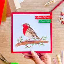 Load image into Gallery viewer, A hand holds a square Christmas greeting card featuring an iris fold robin bird on a branch with holly leaves and red berries, set against a light wood desk background with craft supplies scattered around.
