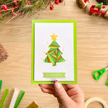 Load image into Gallery viewer, A hand holds a handmade Christmas greeting card featuring an iris fold green tree with a yellow star and the words 'Deck the Halls' on a banner, set against a wooden crafting table background with holiday decorations.
