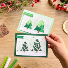 Load image into Gallery viewer, A hand holds a green-bordered Christmas card featuring iris fold green trees, with a second card behind it on a wooden tabletop surrounded by holiday craft supplies.
