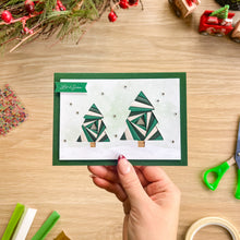 Load image into Gallery viewer, A handmade Christmas greeting card featuring two iris fold green and white Christmas trees, held by a hand over a wooden crafting table with holiday decorations.
