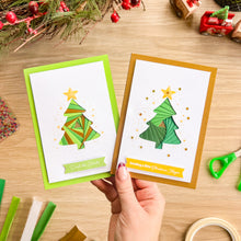 Load image into Gallery viewer, A hand holds two handmade Christmas greeting cards with iris fold tree designs on a wooden tabletop. One card has a bright green border and the other has a metallic gold border.
