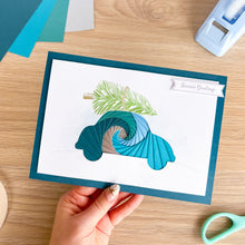 Load image into Gallery viewer, A hand holds a handmade greeting card featuring an iris folding car made from teal, blue, and silver paper, with a green pine branch and a 'Season's Greetings' banner. The card is photographed on a wooden desk with crafting tools like scissors and a tape dispenser visible.
