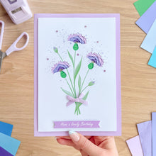 Load image into Gallery viewer, A handmade greeting card with a purple cornflower bouquet design and the text 'Have a lovely Birthday' on a lavender banner, held by a hand over a wooden craft table with card-making tools and colorful paper around it.
