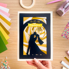 Load image into Gallery viewer, Handmade iris fold card with a couple silhouette and wedding date on a wooden table with craft supplies.
