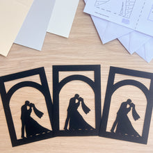 Load image into Gallery viewer, Three black paper cutouts of a couple kissing on a wooden surface with envelopes.
