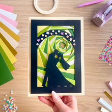Load image into Gallery viewer, Handmade iris fold card with a couple silhouette design held by a hand on a wooden table with craft materials.
