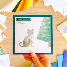 Load image into Gallery viewer, Hand holding a card with a cat and Christmas tree design on a wooden surface with craft materials.
