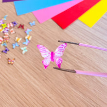 Load image into Gallery viewer, Pink butterfly decorations on a wooden surface with colorful paper strips in the background.
