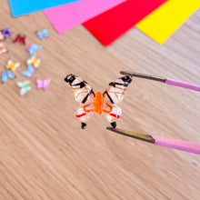 Load image into Gallery viewer, Butterfly-shaped gem being held by tweezers on a wooden surface with colourful paper in the background
