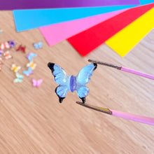 Load image into Gallery viewer, Blue butterfly gem on a wooden surface with colourful paper.
