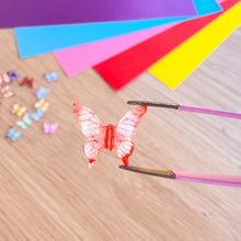 Load image into Gallery viewer, Red butterfly-shaped object being held by tweezers with colourful paper and small butterflies in the background.

