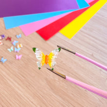 Load image into Gallery viewer, Butterfly-shaped craft item on tweezers with colorful paper in the background

