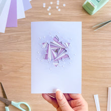 Load image into Gallery viewer, A hand holds a square white card featuring an iris fold snowflake in shades of purple and white, surrounded by small white dots. The card is photographed from above on a light wood desk with crafting tools like scissors, tweezers, and paper sheets visible in the background.
