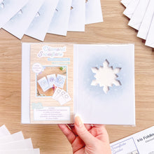 Load image into Gallery viewer, A hand holds a packaged card aperture pack labeled 'Diamond Snowflake' on a light wood tabletop, with the pack contents arranged around it.
