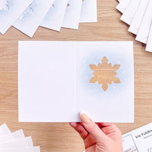 Load image into Gallery viewer, A hand holds an open white folding card featuring a light blue watercolor snowflake design on the right page, set on a light wood tabletop with other cards scattered around.
