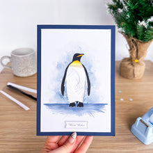 Load image into Gallery viewer, A handmade greeting card featuring a watercolor illustration of an emperor penguin standing on ice, held by a hand over a wooden desk with a mug and small plant in the background.
