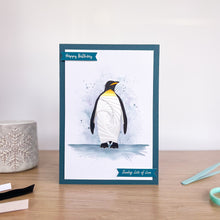 Load image into Gallery viewer, A handmade greeting card featuring an iris fold penguin, standing on a wooden tabletop with a neutral background and craft props nearby.
