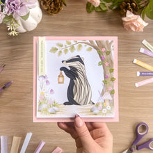 Load image into Gallery viewer, A hand holds a square pink card featuring an iris fold badger holding a lantern under a tree, surrounded by flowers and woodland details. The card is photographed on a wooden tabletop with craft supplies and flowers around it.
