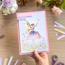 Load image into Gallery viewer, A handmade greeting card featuring a whimsical iris fold fairy sitting on a pink toadstool in a flower field, held by a hand over a wooden craft table surrounded by crafting supplies.
