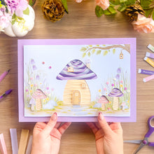 Load image into Gallery viewer, A handmade A4 paper craft card featuring a whimsical toadstool house, held by two hands over a wooden desk decorated with craft supplies and flowers.
