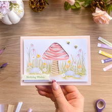Load image into Gallery viewer, A handmade greeting card held by a hand, featuring a pink and white iris fold mushroom with a grassy wildflower background. The card has a 'Birthday Wishes' banner and is displayed on a wooden desk with craft supplies.
