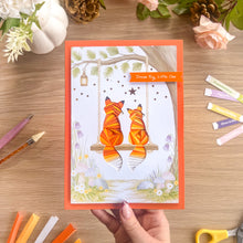 Load image into Gallery viewer, A handmade greeting card featuring two orange and yellow paper-folded foxes sitting on a wooden swing, held by a hand over a wooden desk with craft supplies.
