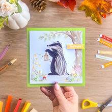 Load image into Gallery viewer, A handmade square greeting card featuring a whimsical iris fold badger holding a lantern in a forest, held by a hand over a wooden desk with autumn-themed craft supplies.
