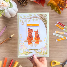 Load image into Gallery viewer, A handmade greeting card featuring two orange foxes sitting on a swing with a 'Happy anniversary' banner, held by a hand over a wooden desk decorated with craft supplies and autumn leaves.
