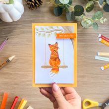Load image into Gallery viewer, A hand holds a greeting card featuring an iris fold orange fox sitting on a swing, with a yellow-orange border and the text 'Wild About You' on a banner. The card is photographed on a wooden tabletop surrounded by craft supplies and greenery.
