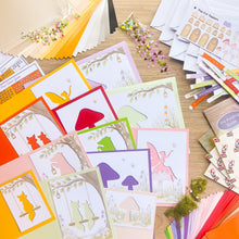 Load image into Gallery viewer, A styled flat-lay of handmade greeting card-making materials featuring whimsical woodland and fairy designs, surrounded by crafting supplies including papers, envelopes, embellishments, and a 'Pop-Cut Toppers' sheet on a light wood background.
