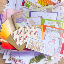 Load image into Gallery viewer, A flat-lay display of a card making kit containing various colourful cardstock sheets, an iris folding guide, pop-cut toppers, glitter, moss, and floral embellishments on a wooden surface.
