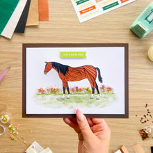 Load image into Gallery viewer, A hand holds a greeting card featuring an iris fold brown and black horse with a green banner that reads 'Love from the Farm' above a watercolor field of flowers. The card is photographed on a wooden craft table surrounded by crafting supplies.
