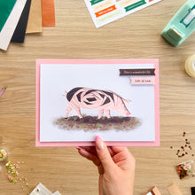 Load image into Gallery viewer, A hand holds a handmade iris fold greeting card featuring a pink and black patterned pig on a white background with a pink border, set against a wooden desk with crafting supplies.
