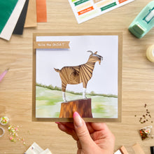 Load image into Gallery viewer, A hand holds a square greeting card featuring an iris fold goat standing on a tree stump with the text 'You're the GOAT'. The card is photographed on a wooden desk surrounded by craft supplies.
