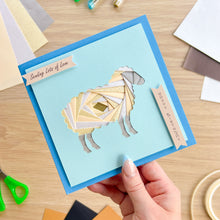 Load image into Gallery viewer, A handmade square greeting card held by a hand, featuring an iris fold sheep design. The card has a medium blue border and small ribbon banners reading 'Sending Lots of Love' and 'Happy Birthday'.

