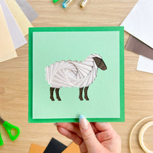 Load image into Gallery viewer, A handmade greeting card featuring an iris folded sheep on a mint green background, held by a hand over a wooden craft table with colorful cardstock and scissors.

