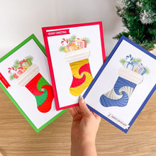 Load image into Gallery viewer, Three Christmas cards with stockings and presents on a wooden surface.
