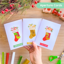 Load image into Gallery viewer, Three Christmas-stocking themed greeting cards are held in a hand over a wooden tabletop decorated with pine branches, red berries, and craft scissors. The cards feature colourful illustrated iris fold Christmas stockings with gift boxes and holiday messages.
