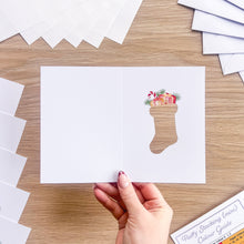 Load image into Gallery viewer, A hand holds a white greeting card with a Christmas stocking cut-out, filled with small illustrated gifts and evergreen branches, on a light wood tabletop. More white cards are fanned out around it.
