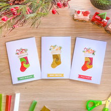 Load image into Gallery viewer, Three Christmas greeting cards with colourful iris fold stocking are arranged on a wooden tabletop with holiday-themed props including pine branches, berries, scissors, and toy trains.
