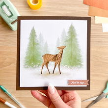 Load image into Gallery viewer, Card with a deer design held by a hand on a wooden surface with stationery items.

