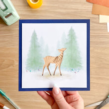 Load image into Gallery viewer, A hand holds a square greeting card featuring an iris fold deer in front of watercolor pine trees. The card has a navy blue border and is photographed on a wooden desk with crafting tools like scissors, tape, and gel pens visible around it.
