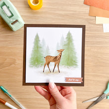 Load image into Gallery viewer, A hand holds a square greeting card featuring an illustration of a brown deer in a snowy pine forest. The card has a dark brown border and a small banner reading 'Just to say.
