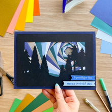 Load image into Gallery viewer, A handmade layered card with a forest silhouette and geometric sunburst pattern in shades of blue, gray, and white, held by a hand over a wooden table with craft paper and scissors.
