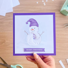 Load image into Gallery viewer, A hand holds a square Christmas greeting card with a purple border and a white iris fold snowman, with 'MERRY CHRISTMAS' on a purple banner. The card is photographed on a light wood desk with scissors and other crafting items nearby.
