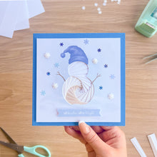 Load image into Gallery viewer, A handmade square greeting card held by a hand, featuring a winter-themed design with an iris fold snowman, a blue hat, snowflake decorations, and a blue border. The card is photographed on a wooden desk with crafting tools like scissors and tape visible in the background.
