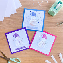 Load image into Gallery viewer, Three handmade square Christmas greeting cards with iris fold snowman illustrations are arranged on a wooden tabletop with scissors, paper, and a tape dispenser nearby.
