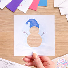Load image into Gallery viewer, A hand holds a square white paper craft cut-out featuring a snowman with a blue hat, shown over a light wood table with colorful paper, envelopes and an Iris Folding Pattern.
