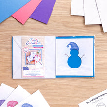 Load image into Gallery viewer, A flat-lay of a 'Frosty Snowman' iris folding card aperture pack on a light wood tabletop. The kit contents surround the pack.
